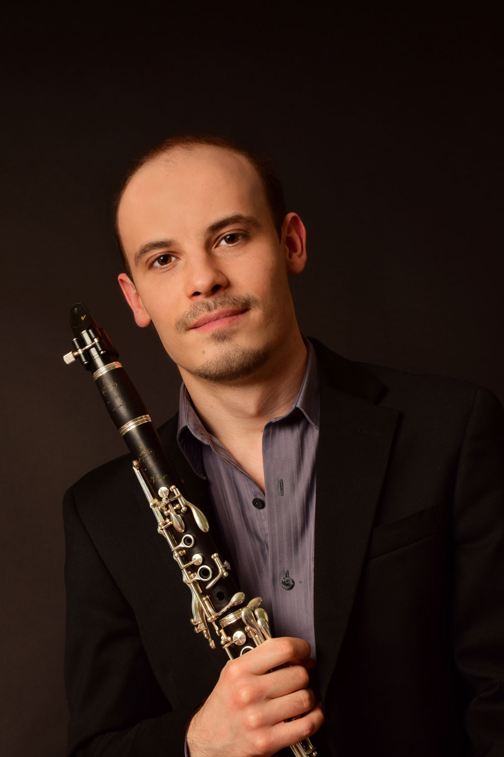Portrait of a man in a black blazer holding a clarinet, looking at the camera against a dark backdrop.
