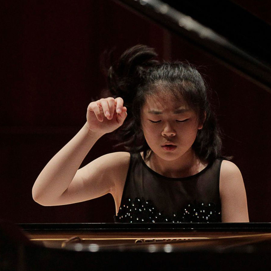 Young girl in a black beaded dress concentrates at a piano, her right hand raised mid-performance.