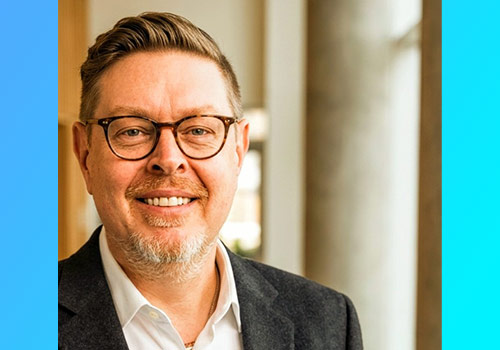 Smiling man in a dark blazer and glasses, indoors near a columned wall.
