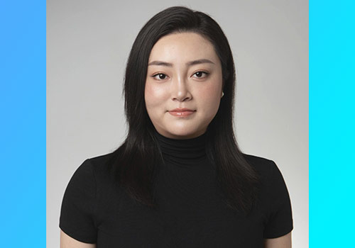 Portrait of a woman with straight black hair in a black turtleneck, neutral expression, against a light gray backdrop with blue side panels behind.