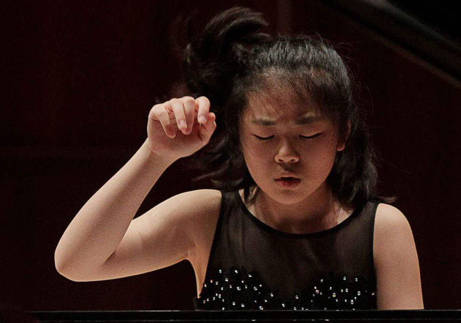 Young girl in a black beaded dress concentrates at a piano, her right hand raised mid-performance.