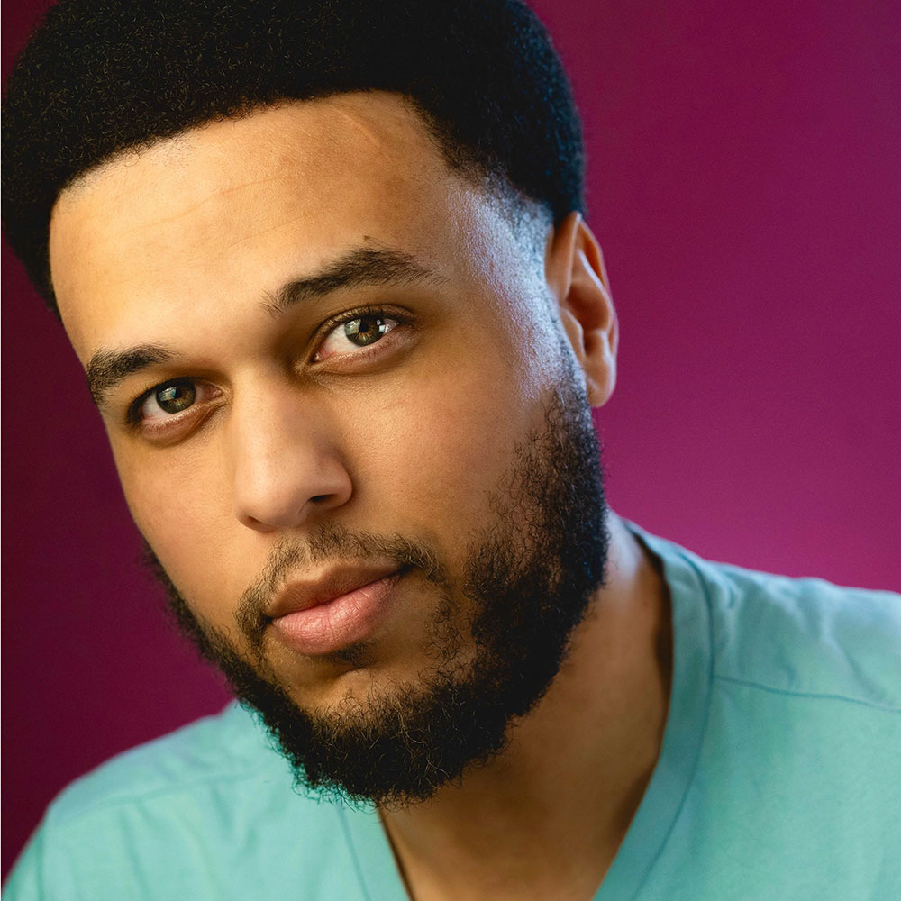 Close-up portrait of a man with a beard wearing a light teal shirt against a magenta background.