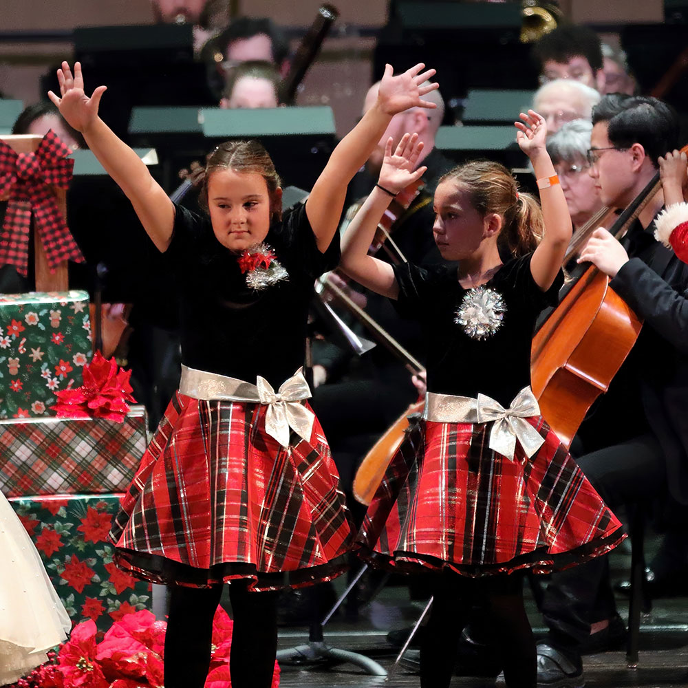 Two young girls in black tops and red plaid skirts dance with arms raised on a holiday stage, an orchestra in the background.