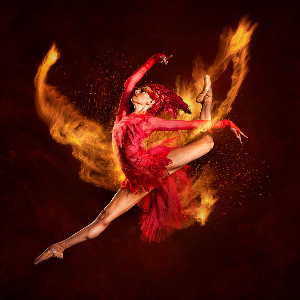 Red-costumed dancer performing a high leap with flames swirling around her against a dark background.